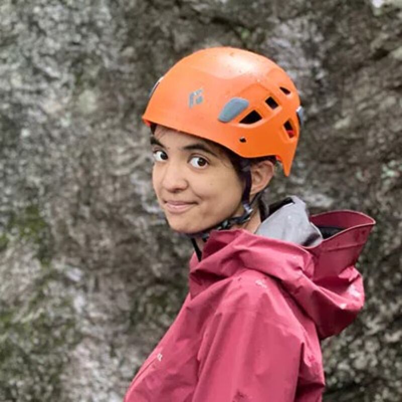 The image shows a person wearing an orange helmet and a red jacket. The person is looking at the camera and smiling. The background is a blurry rock face. The person appears to be outdoors, possibly hiking or climbing.